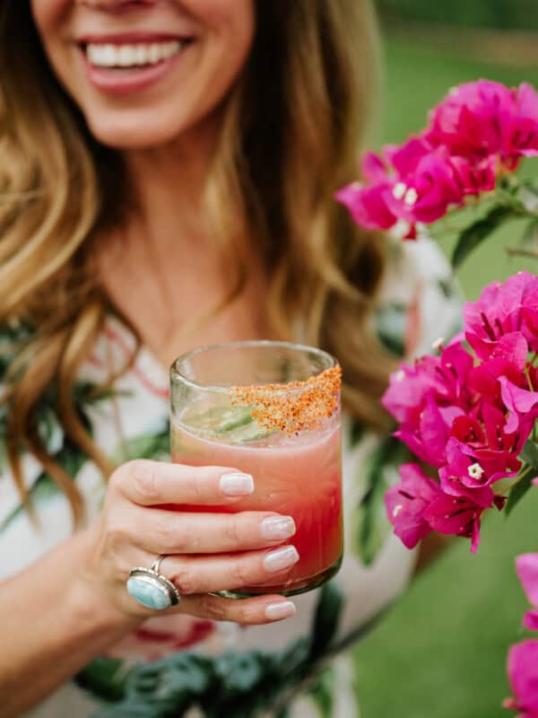 Latina blogger holding a tajin-rimmed jalapeno watermelon margarita near a flower bush.