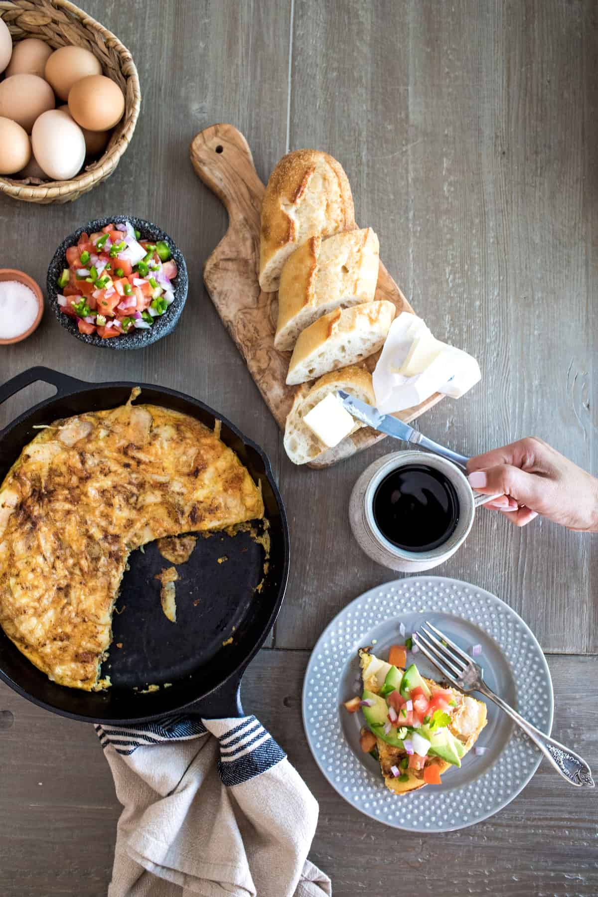 flat lay shot of a breakfast table with a cast iron skillet filled with chorizo potato frittata with a few slices taken out, one slice of frittata on a plate topped with garnishes, and a woman's hand grabbing a cup of coffee.