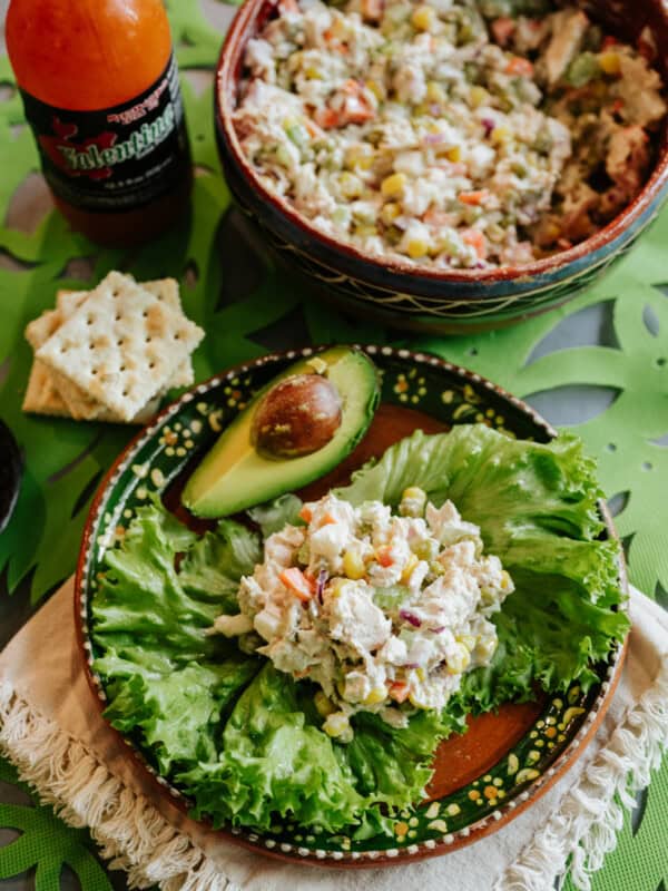 table set with a bowl of ensalada de atun, a bottle of valentina hot sauce, and a plate with a lettuce leaf filled with Mexican tuna salad, a quarter of an avocado on the side, and a handful of saltines.