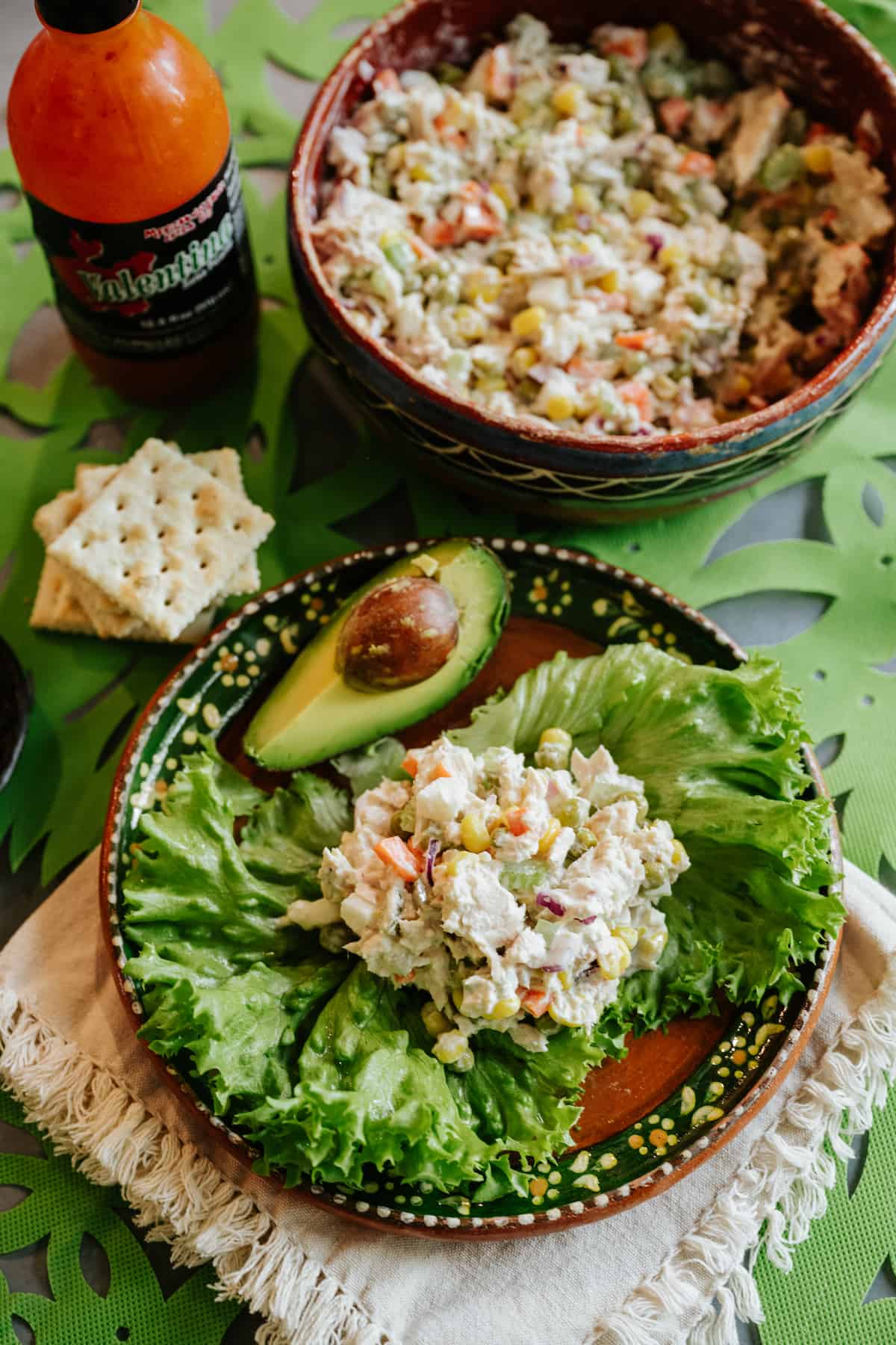 table set with a bowl of ensalada de atun, a bottle of valentina hot sauce, and a plate with a lettuce leaf filled with Mexican tuna salad, a quarter of an avocado on the side, and a handful of saltines. 