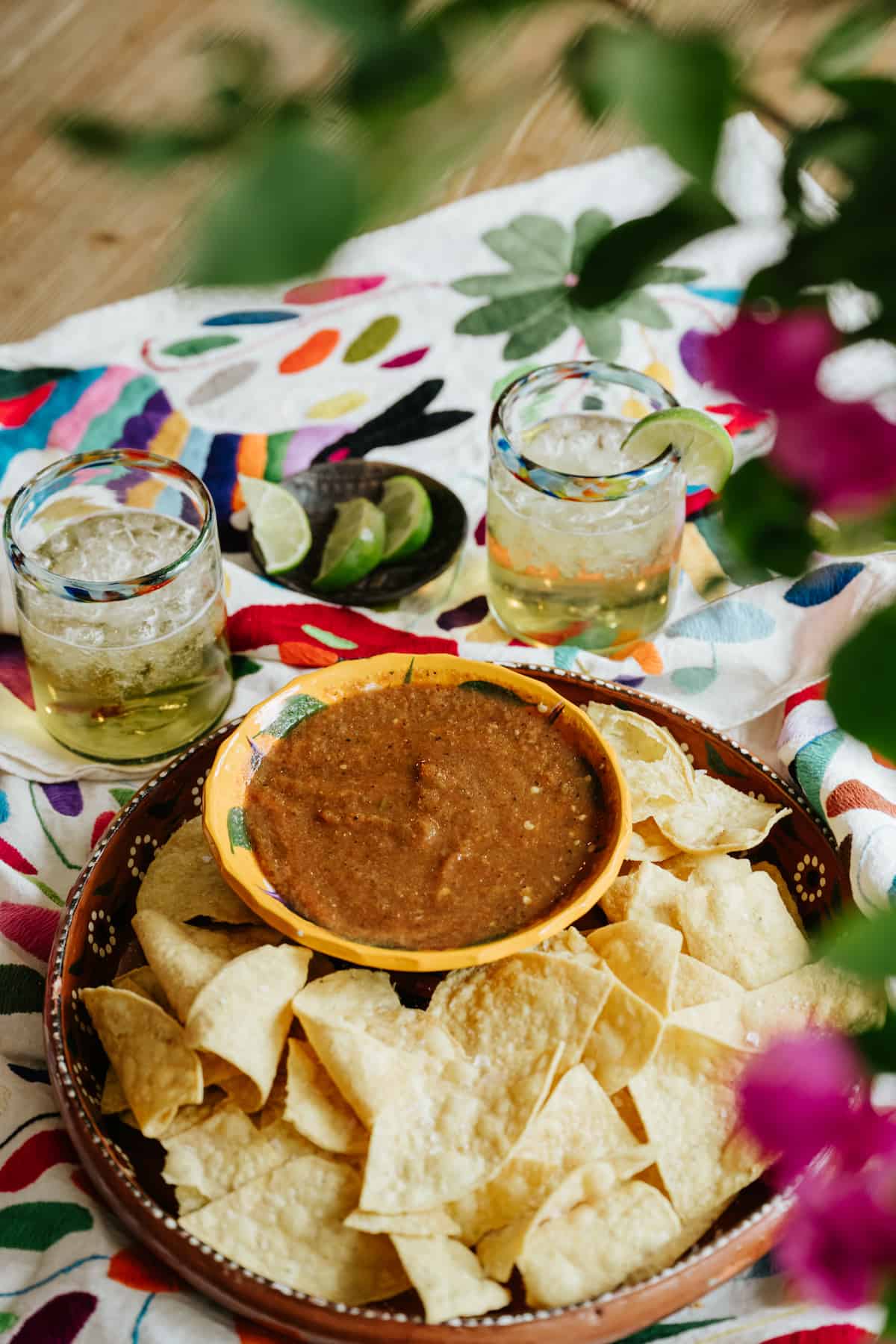 outdoor table set with a colorful tablecloth, two icy glasses of cocktails, and a serving platter of tortilla chips with a bowl of cantina-style salsa roja. 