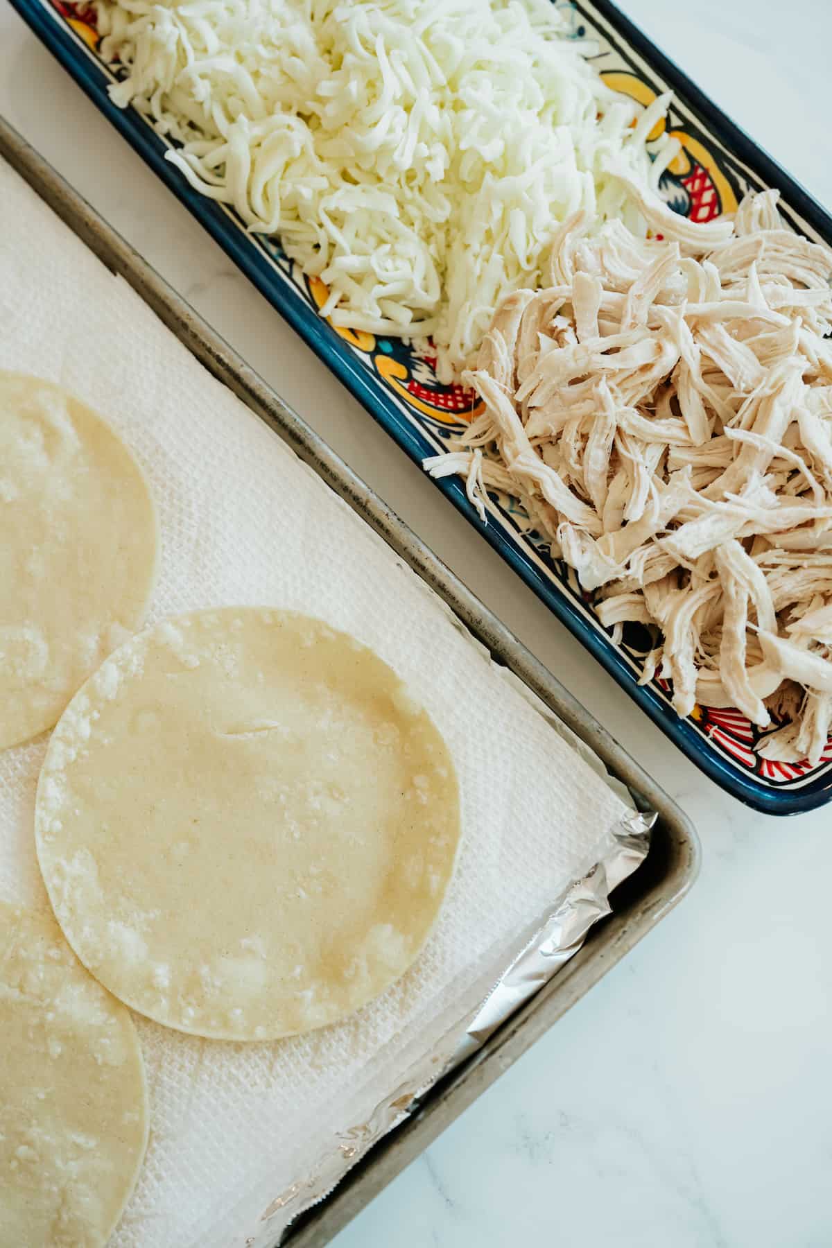 shredded poached chicken breast and cheese on a rectangular plate next to a sheet pan with fried corn tortillas draining on paper towels. 