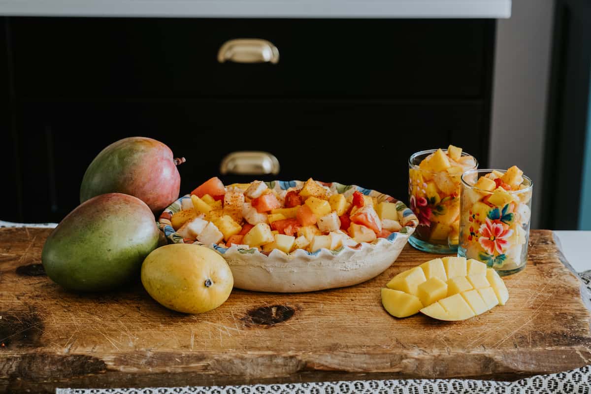 side on shot of a bowl and two glasses filled with Mexican fruit salad with Tajin on a wooden cutting board with mangoes. 