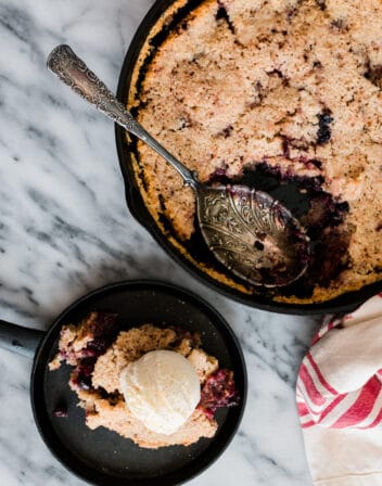 overhead shot of a large skillet filled with mexican chocolate and mixed berry cobbler with one serving put on a black dessert plate and topped with a scoop of vanilla ice cream.