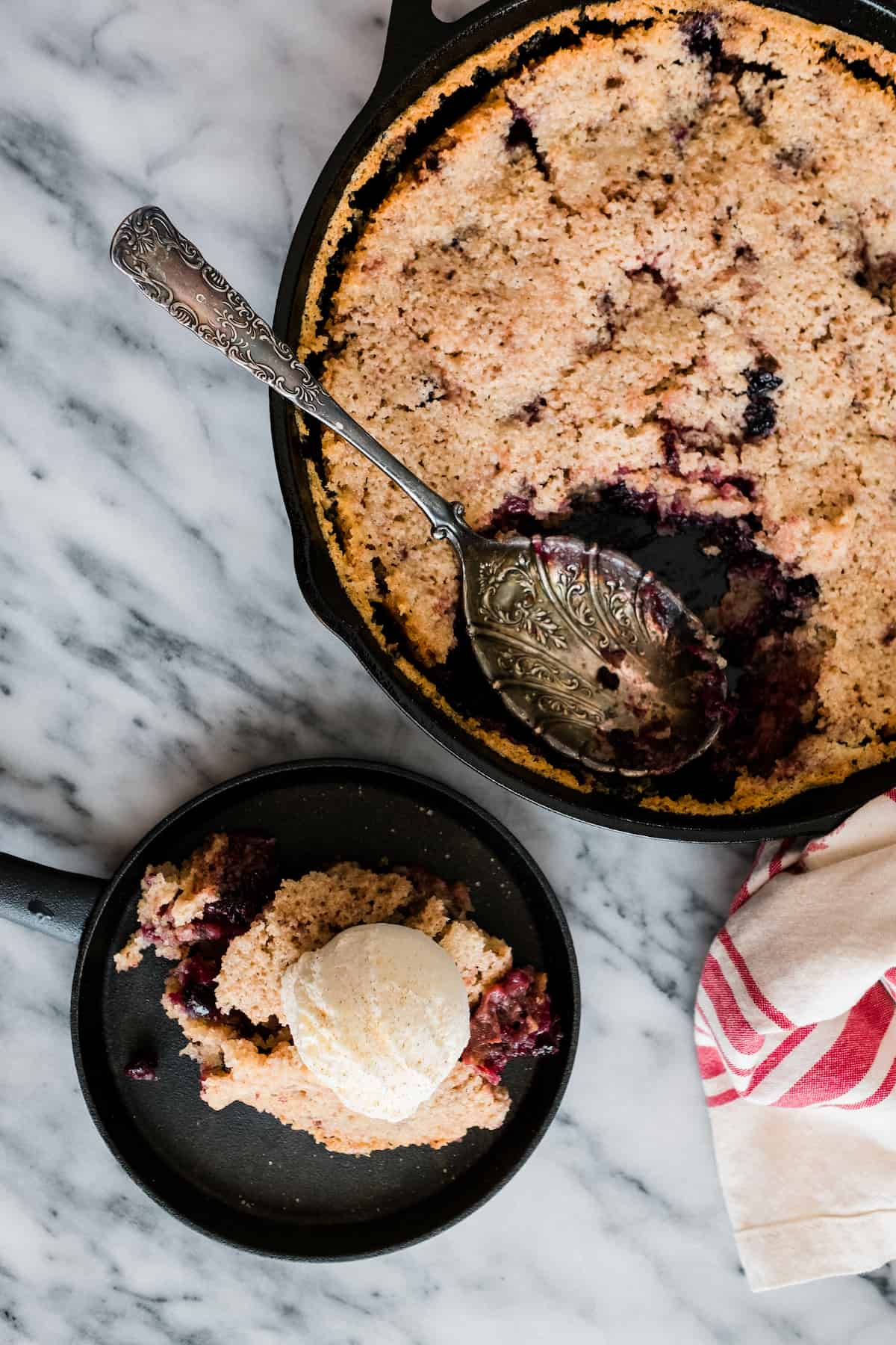 overhead shot of a large skillet filled with mexican chocolate and mixed berry cobbler with one serving put on a black dessert plate and topped with a scoop of vanilla ice cream. 