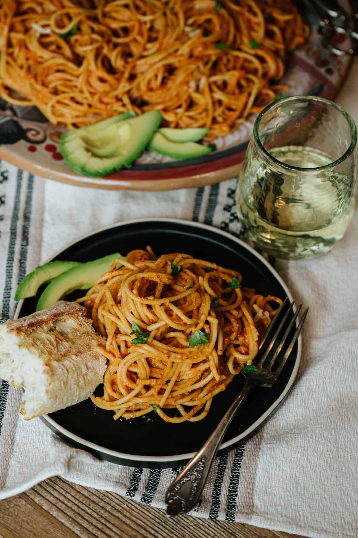black plate with mexican pasta in chipotle cream sauce with a side of crusty bread and some sliced avocado. 