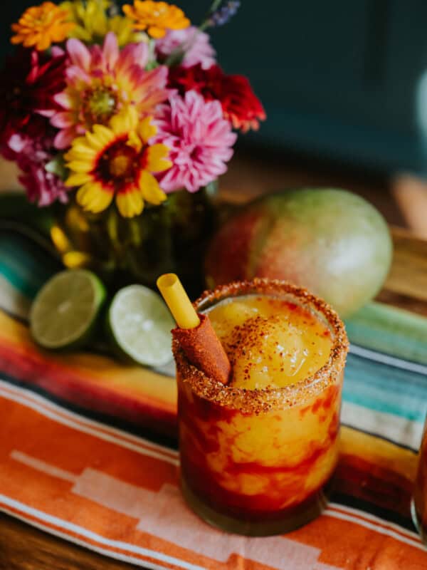 45 degree angle shot of a rocks glass rimmed in tajin and filled with mangonada swirled with chamoy and garnished with a tamarind straw.