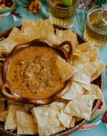 Salsa Borracha (a.k.a. "Drunk Salsa") in a brown pottery bowl surrounded with homemade corn tortilla chips on a turquoise tablecloth with beers in glasses in the background.