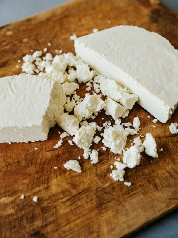 halved block of queso fresco on a wooden cutting board with some of it crumbled.