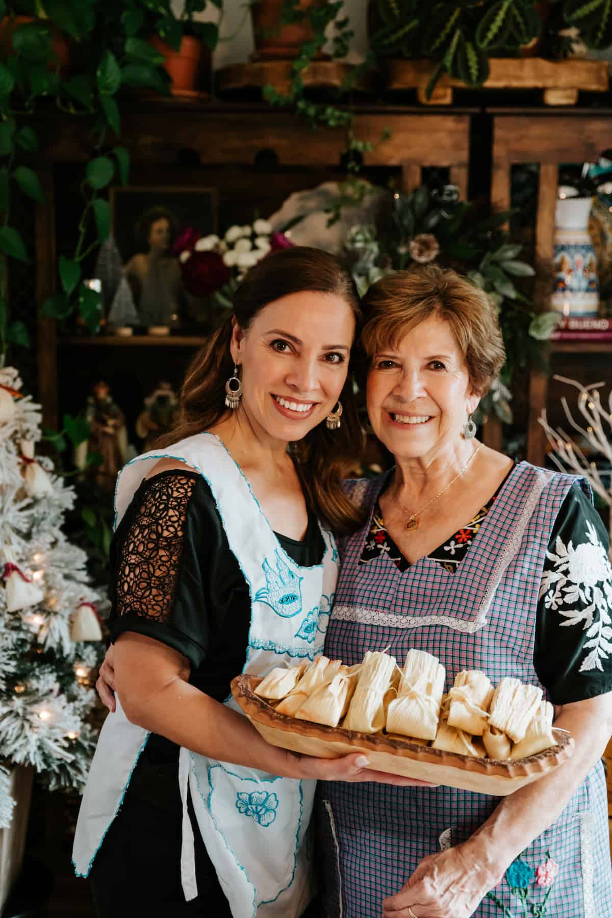 Latina food blogger Yvette Marquez with her mother, both wearing Mexican aprons and holding a tray of homemade wrapped tamales in front of a Christmas tree decorated with mini tamale-shaped ornaments.