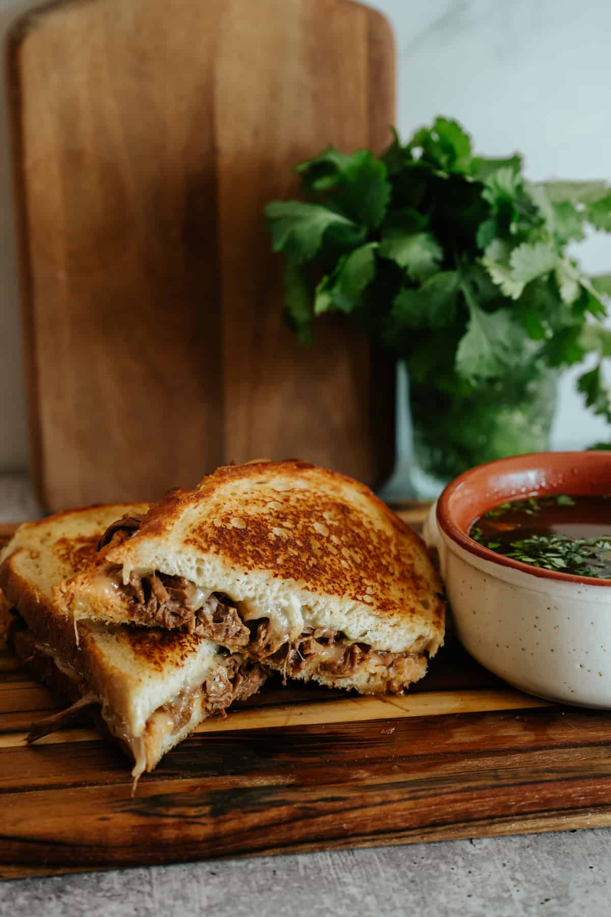 golden, gooey birria grilled cheese sandwich on a rectangular wooden plate next to a bowl of consomme.