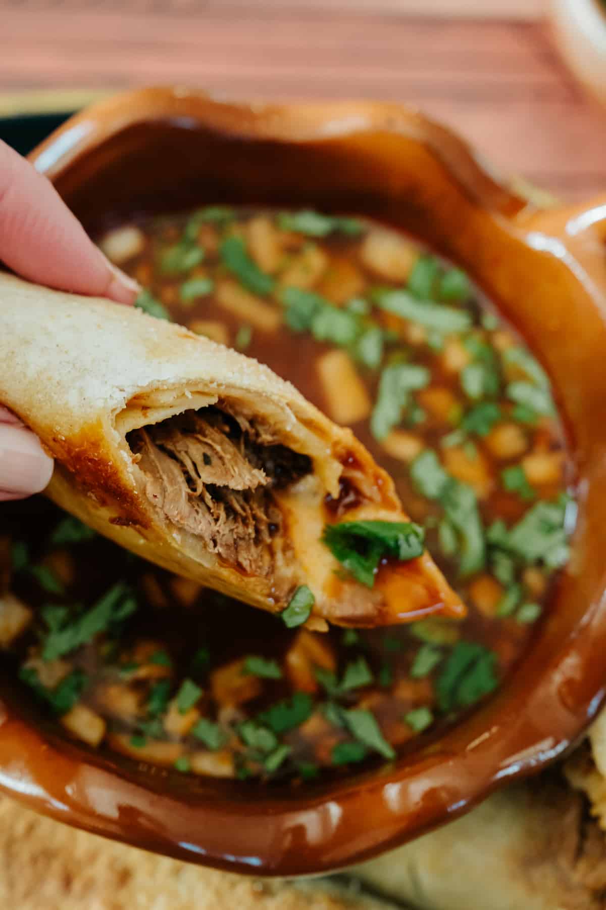 hand holding a baked birria beef flauta above a bowl of the dipping broth.