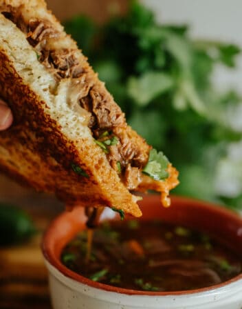 closeup shot of a hand holding a birria grilled cheese sandwich that has been dipped in the consomme.
