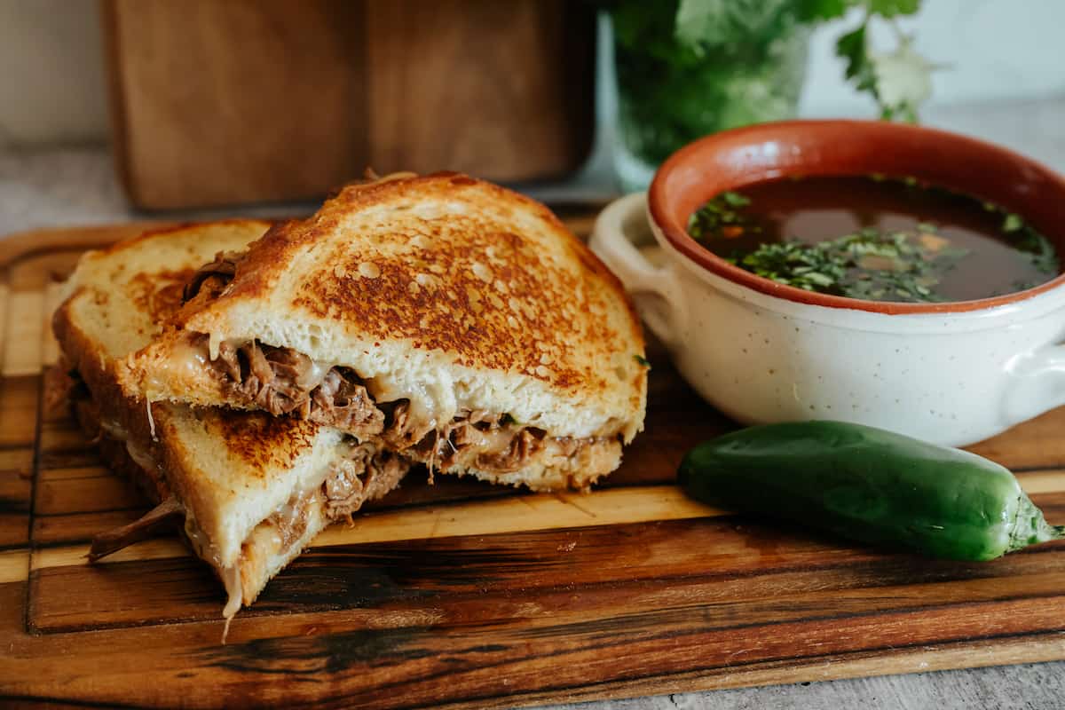 wooden cutting board with two halves of a birria grilled cheese sandwich next to a jalapeño and a bowl of consomme. 