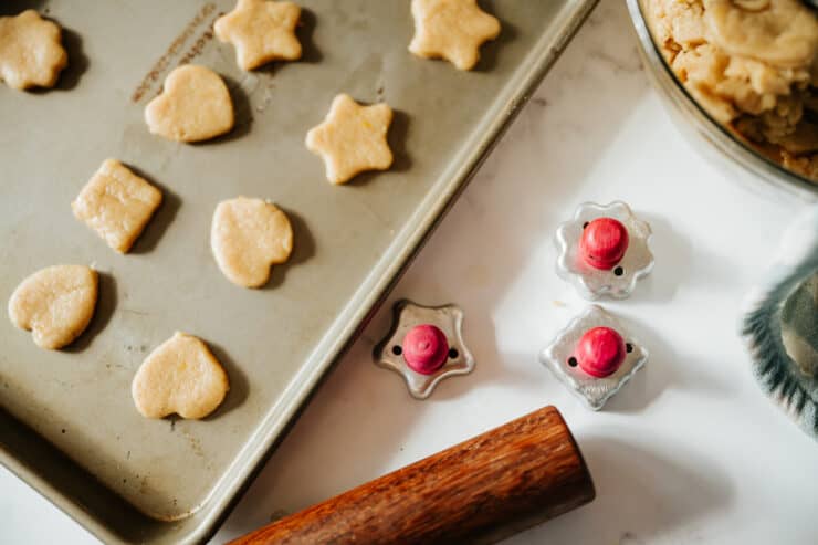 Unbaked biscochos in heart, star, and flower shapes on a baking sheet next to vintage-style cookie press molds and a bowl of dough.