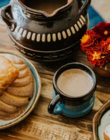 wooden table with a hand thrown mug filled with champurrado next to a platter of pan dulce.