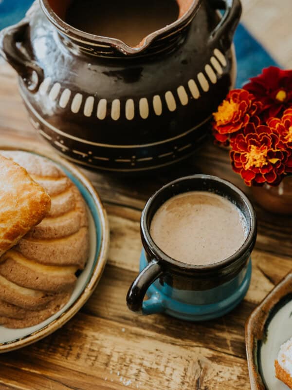 wooden table with a hand thrown mug filled with champurrado next to a platter of pan dulce.