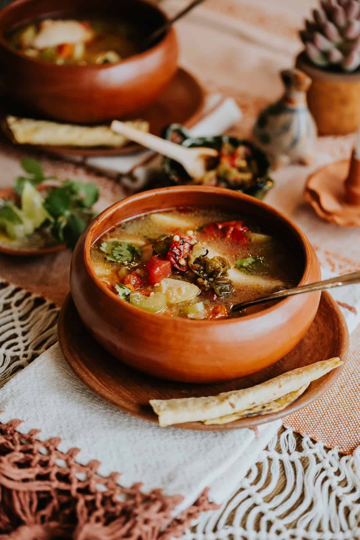 hero shot of a bowl of caldo de pollo Mexican chicken soup on a table with a plate of lime wedges, cilantro, roasted green chiles, and fresh corn tortillas. 