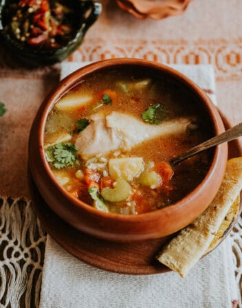 earthenware barre bowl of caldo de pollo soup on a matching plate with a homemade corn tortilla.