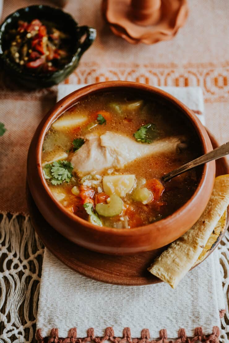 earthenware barre bowl of caldo de pollo soup on a matching plate with a homemade corn tortilla.