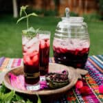 Two tall glasses of agua de jamaica (Mexican hibiscus tea) with ice and fresh mint on a colorful outdoor table, with a glass beverage jar and ladle in the background.