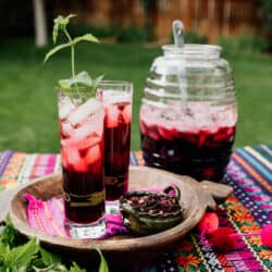 Two tall glasses of agua de jamaica (Mexican hibiscus tea) with ice and fresh mint on a colorful outdoor table, with a glass beverage jar and ladle in the background.