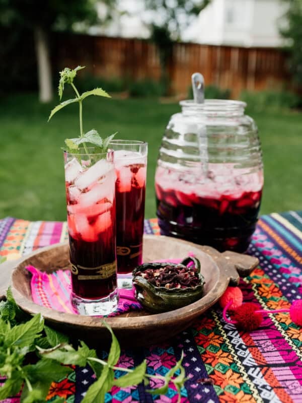 two tall clear collins glasses filled with agua de jamaica, garnished with fresh mint on a colorful outdoor table with a beehive jar in the background with the rest of the hibiscus tea and a ladle.