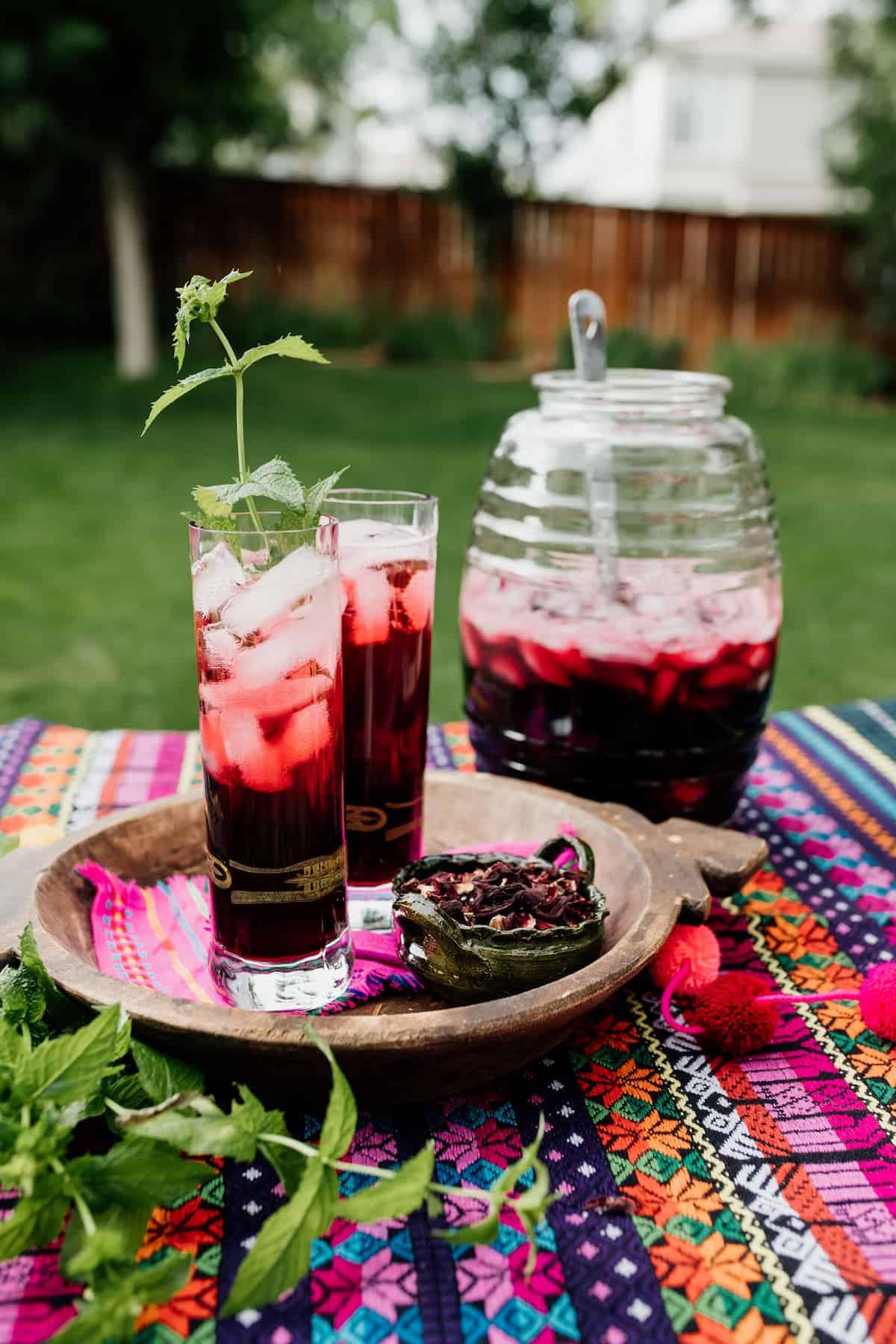 two tall clear collins glasses filled with agua de jamaica, garnished with fresh mint on a colorful outdoor table with a beehive jar in the background with the rest of the hibiscus tea and a ladle. 