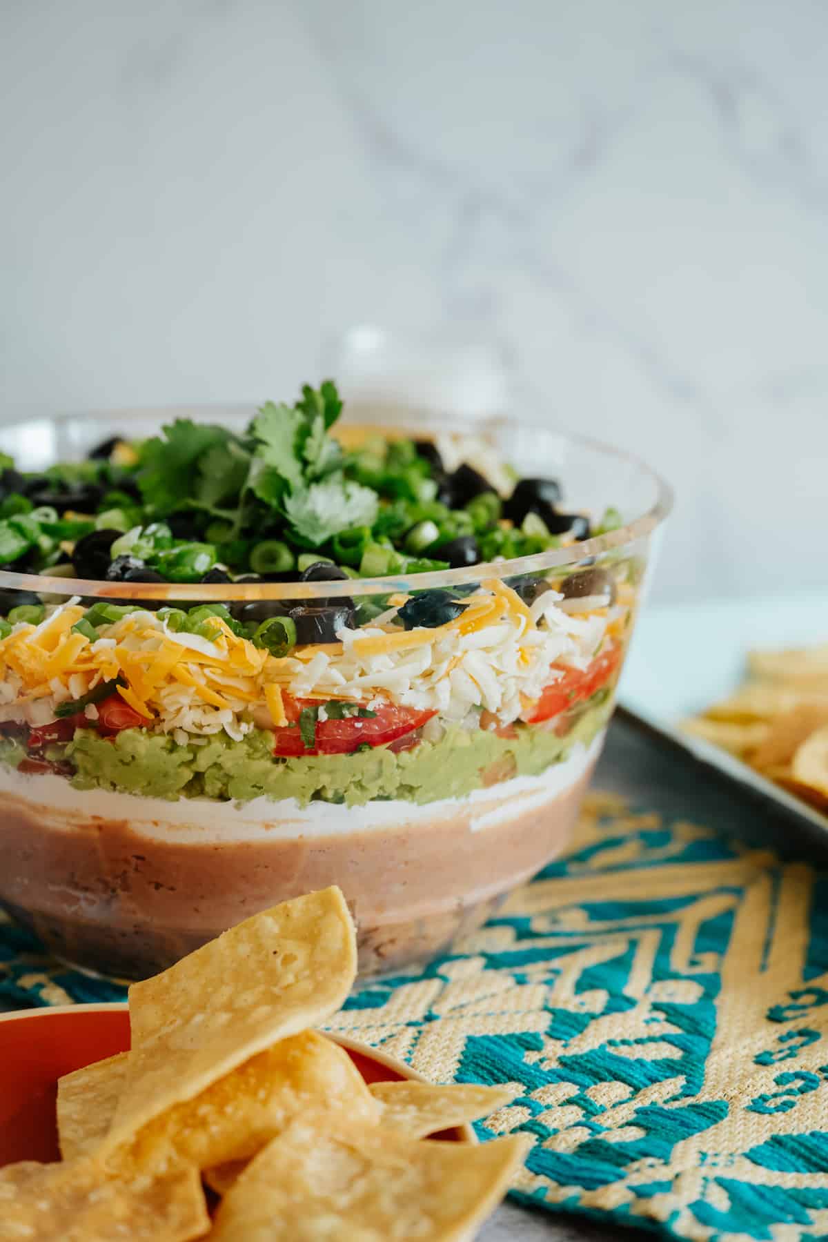 7 layer dip in a bowl on a Mexican textile with chips scattered.