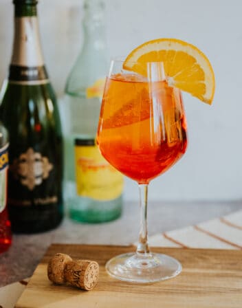 hero shot of a stemmed wine glass filled with an Aperol spritz on ice garnished with an orange round in front of a cava and topo chico bottle.