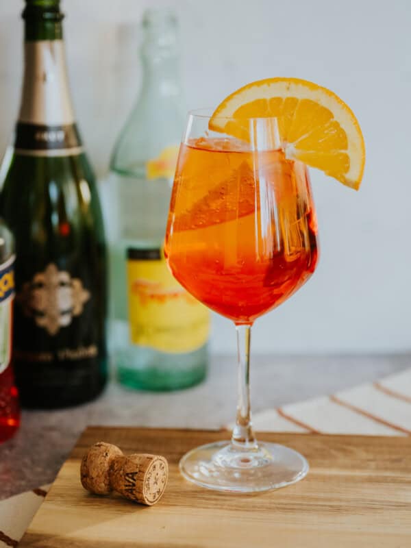 hero shot of a stemmed wine glass filled with an Aperol spritz on ice garnished with an orange round in front of a cava and topo chico bottle.