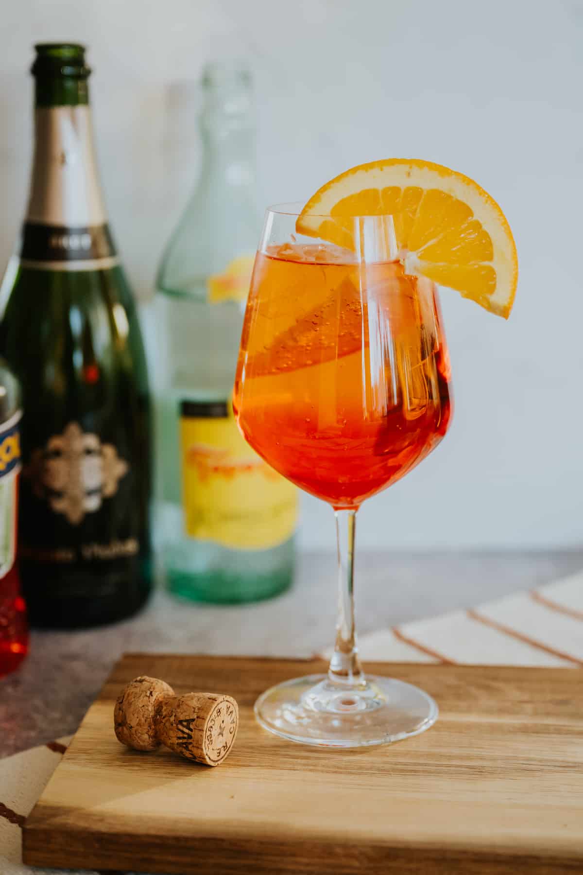 hero shot of a stemmed wine glass filled with an Aperol spritz on ice garnished with an orange round in front of a cava and topo chico bottle. 