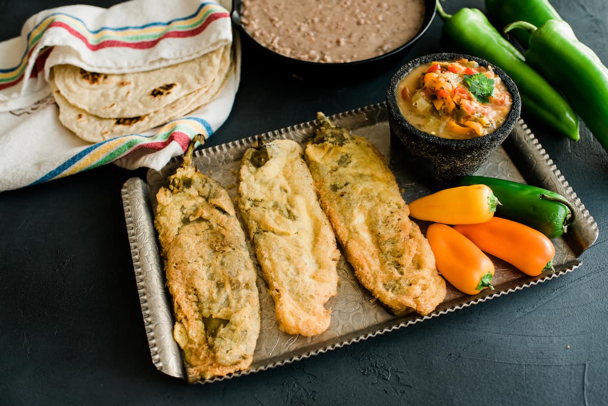 3 fried chiles rellenos capeados on a silver serving platter next to smaller whole green and orange peppers, a small molcajete of roasted pepper salsa, a bowl of refried beans and a towel of warmed flour tortillas. 