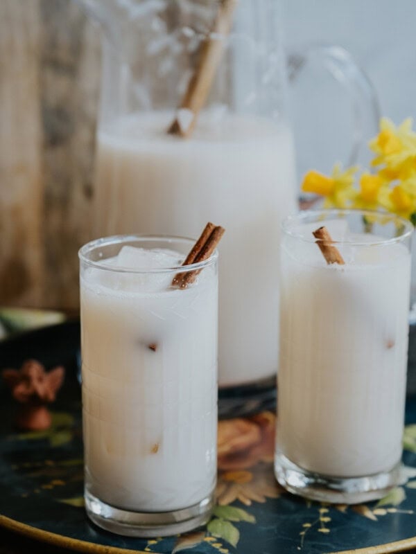 two tall glasses of horchata on ice with a pitcher in the background.