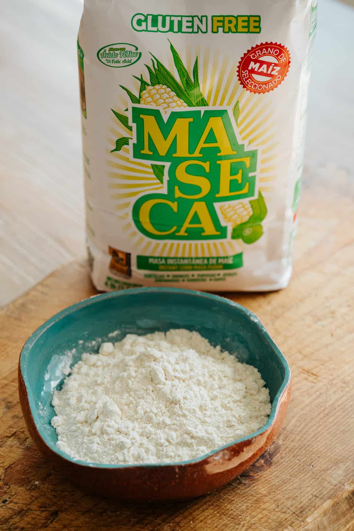 45 degree angle shot of a turquoise bowl filled with masa harina on a wooden cutting board with a bag of Maseca brand masa harina in the background. 