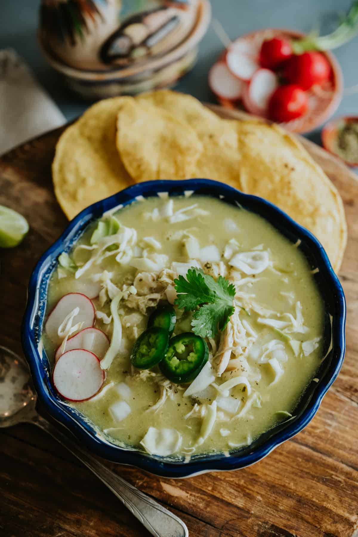 navy blue bowl filled with chicken pozole verde topped with jalapeno and radish slices, shredded lettuce, and fresh cilantro leaves.