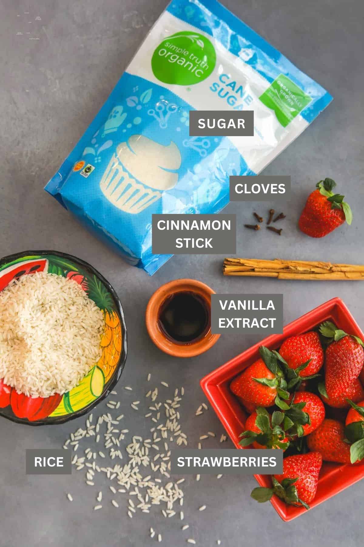 flat lay shot of rice, strawberries, and spices needed to make strawberry horchata (agua de horchata de fresas) labeled on a grey table.