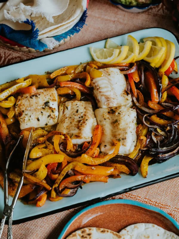 flat lay shot of a rectangular pale blue serving platter with cod and fajita veggies next to a plate of warm flour tortillas for making sheet pan fish fajitas.