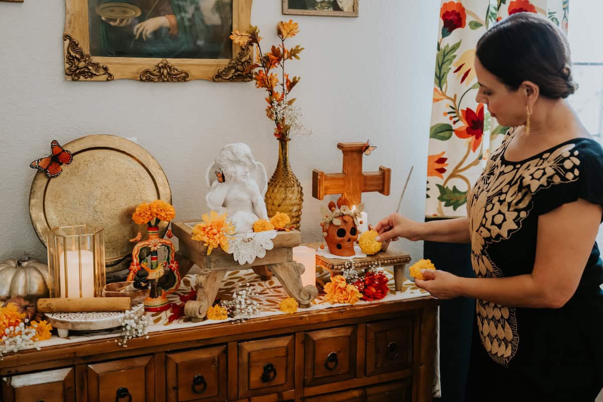 Yvette Marquez, blogger behind Muy Bueno, standing in front of an ofrenda placing a yellow marigold next to a cross; there is a wooden Mexican palote (rolling pin) to her left.