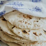 stack of homemade flour tortillas loosely wrapped in a blue and white flour sack towel on a wooden pedestal next to a Mexican wooden rolling pin.