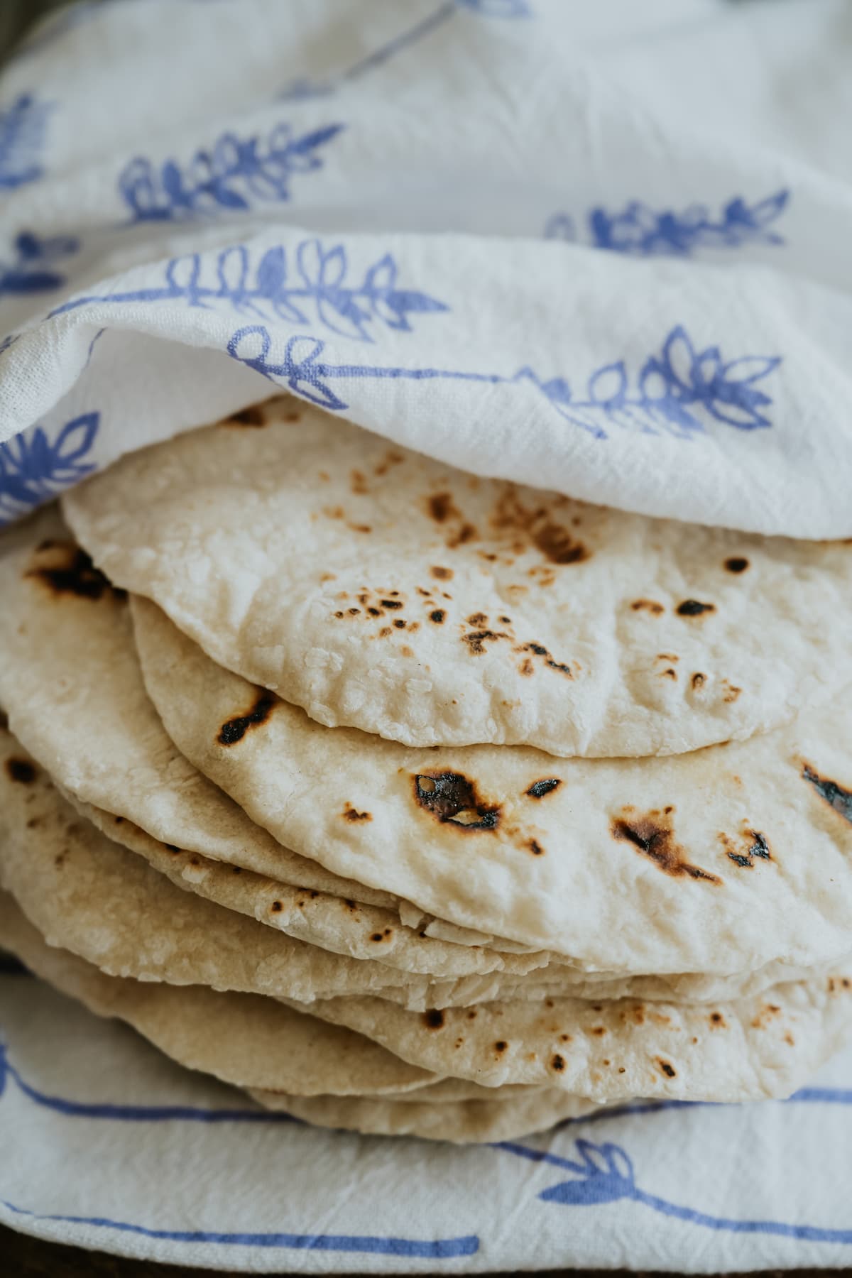 closeup shot of a stack of freshly made flour tortillas wrapped in a clean white kitchen towel with a blue floral pattern. 
