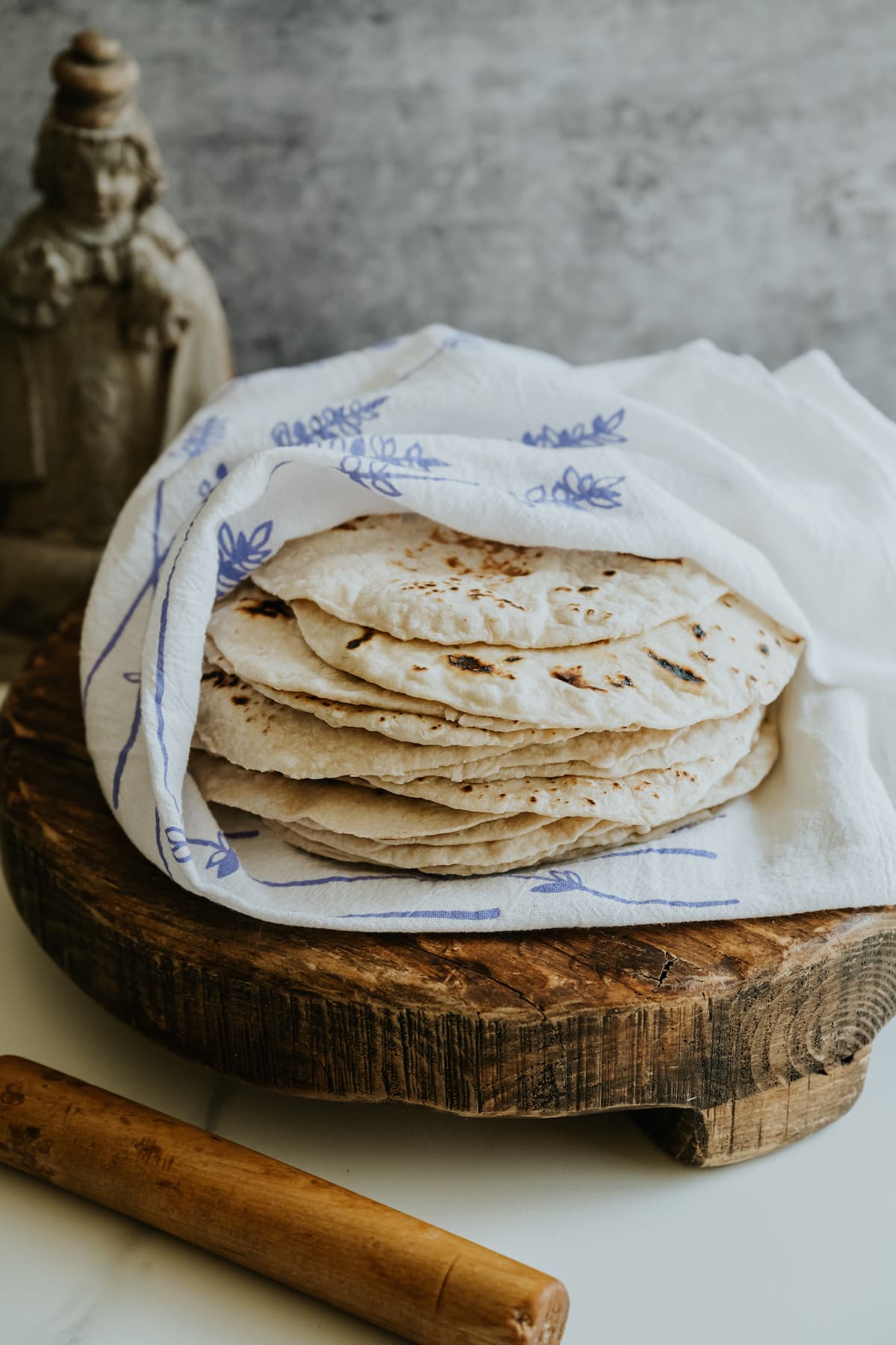 stack of homemade flour tortillas loosely wrapped in a blue and white flour sack towel on a wooden pedestal next to a Mexican wooden rolling pin. 