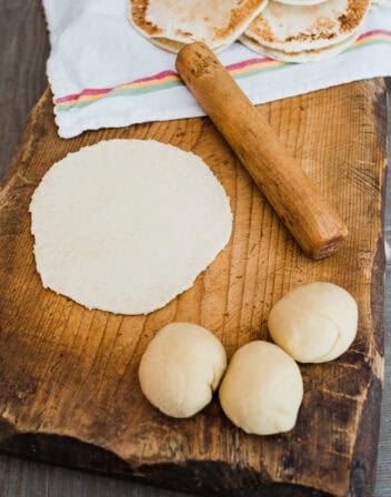 wooden cutting board with balls of dough and a rolled out tortilla next to a matching wooden palote.