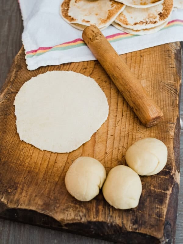 wooden cutting board with balls of dough and a rolled out tortilla next to a matching wooden palote.