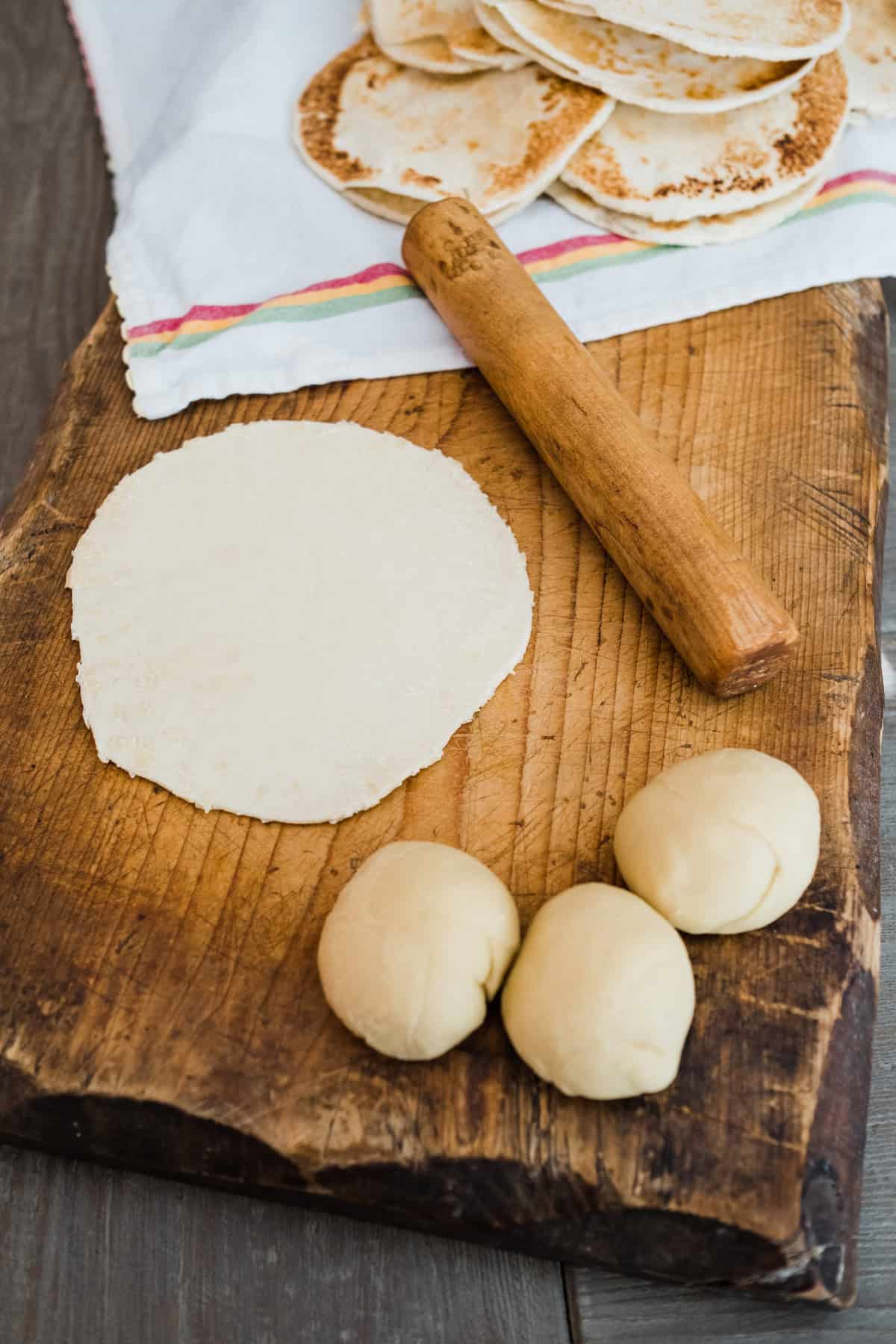 wooden cutting board with balls of dough and a rolled out tortilla next to a matching wooden palote. 