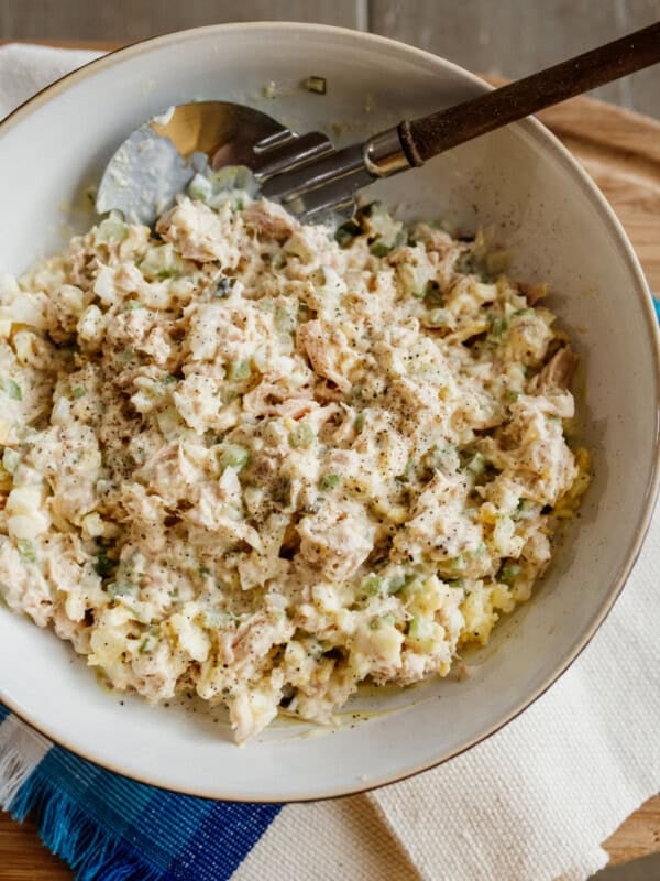 tuna salad made with eggs in a serving bowl with a wooden handled serving spoon on a blue and white woven cloth.