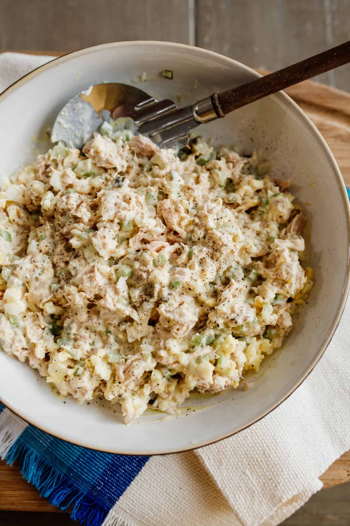 tuna salad made with eggs in a serving bowl with a wooden handled serving spoon on a blue and white woven cloth.