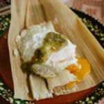 green chile tamal on top of its corn husk wrapper on top of a terracotta plate served with a runny fried egg and a dollop of salsa verde.