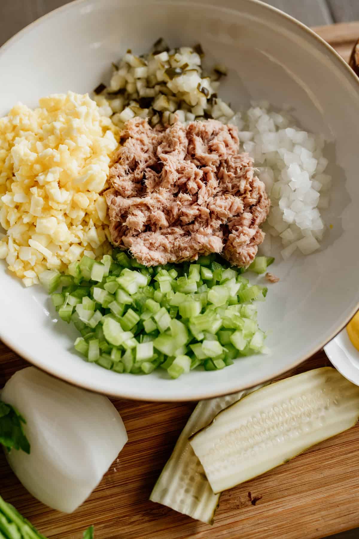 close up of diced celery chopped hard boiled eggs onion pickles and canned tuna in a white bowl before mixing into tuna egg salad. 