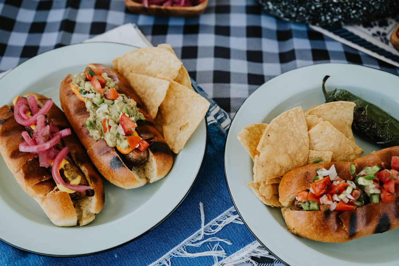 two pale blue metal camping plates with Mexican-adorned bacon-wrapped hot dogs (including pickled red onions, chunky guacamole, fresh pico de gallo, roasted jalapeños and crema mexicana atop a black and white gingham tablecloth. 